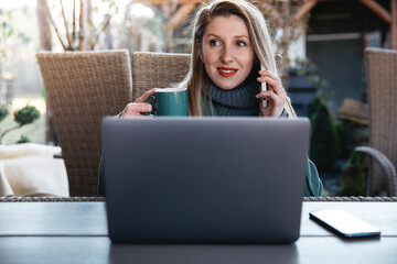 Naklejka premium Beautiful young woman with laptop and talking on cell phone and smiling. Female using cell mobile phone while sitting at home garden outdoors.
