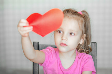 a little girl looks at a red heart cut out of colored paper, preparing for the holiday Mother's Day, Valentine's Day. DIY holiday card with a red paper heart, a symbol of love