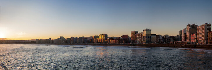 Gijón at dawn over the beach of San Lorenzo. Panoramic view of Gijón with the sun reflecting on the buildings of the city and the sea. The beach of San Lorenzo seen from the church of San Pedro.