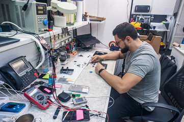 Electronics repair shop, a repairman is surrounded by tools and equipment. A technician repairs, cleans, controls a smartphone. Workplace top view, close-up.