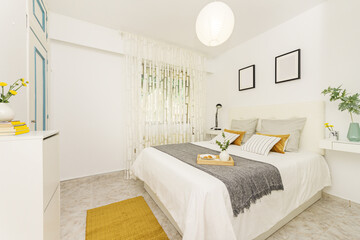Bedroom with a double bed with a white leather upholstered headboard with various types of cushions and a wooden tray with a vase and rolls in a white bowl, yellow rugs and a window with curtains