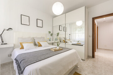 Fototapeta na wymiar Bedroom with a double bed with a white leather upholstered headboard with cushions and a wardrobe with sliding mirror doors and white wood