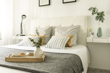 Bedroom with a double bed with a white leather upholstered headboard with cushions of various types and a wooden tray with a vase and rolls in a white bowl