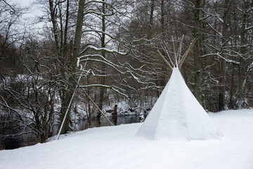 Tipi tent covered with snow next to a river by the forest. Winter glamping outdoors, camp site