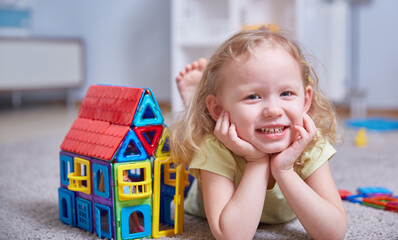 Cute curly girl next to a house made of a magnetic constructor at home on the carpet in the children's room.