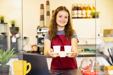 Portrait of a young beautiful woman in red apron, keeps white cup in hand, smiling, looking at camera, on a coffee shop background.