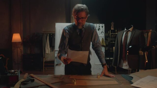 Male mature tailor in luxury designer atelier or tailoring studio. He works with sketch of future garment on table with fabric and tools. Mannequin with tailored suit. Fashion and hand craft concept.