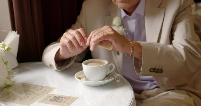mature man groom breaks a sugar bag and pours sugar into a cup of cappuccino coffee in a restaurant. elderly handsome old man sugaring coffee at the table