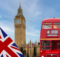 Old traditional vintage red London Bus driving by Big Ben and the Palace of Westminster in London, England with the flag of the United Kingdom waving in the foreground. Blue sky background.