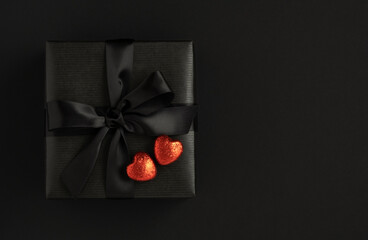 Black gift box with a black bow and two red shiny hearts on a black background. Present for Valentine's Day. Space for your text