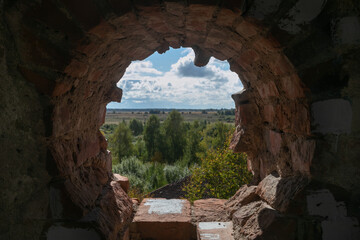 A large round hole in the shabby brick wall. View through a cement pipe to the countryside. In the distance, a blurred landscape: forest, cloudy sky, outlines of buildings. 