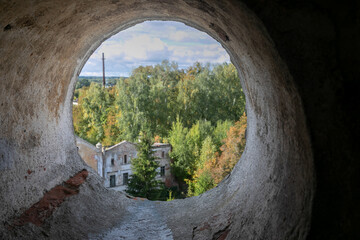 A large round hole in the shabby brick wall. View through a cement pipe to the countryside. In the distance, a blurred landscape: forest, cloudy sky, outlines of buildings. 