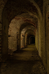 A long dark dimly tunnel of an abandoned military bunker or bomb shelter with broken red brick walls. The arched ceiling is illuminated by daylight from the outside. The floor is dirty and dusty
