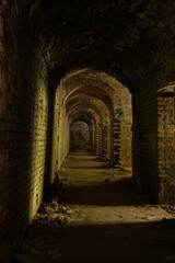 A long dark dimly tunnel of an abandoned military bunker or bomb shelter with broken red brick walls. The arched ceiling is illuminated by daylight from the outside. The floor is dirty and dusty