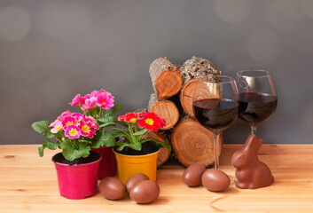 Easter concept. Primula flowers, glasses of red wine, chocolate easter eggs, chocolate bunny and  pile of firewood on wooden table.