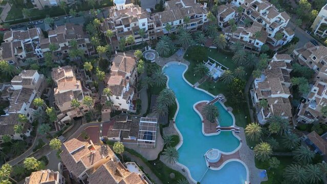 Top-down aerial view of residential neighbourhood showing condos  and multi-story  buildings with a large public turquoise pool among the palm trees. Torrevieja Spain, sunset time, drone footage