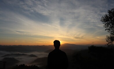 A man facing in front of the sun is rising from a back mountain and there has a sea of fog too.