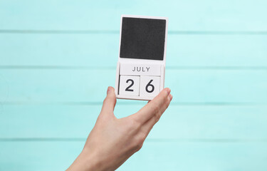 Woman's hand holds wooden calendar with date july 26 on blue wooden background