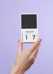 Woman's hand holds wooden calendar with date august 17 on pastel purple background