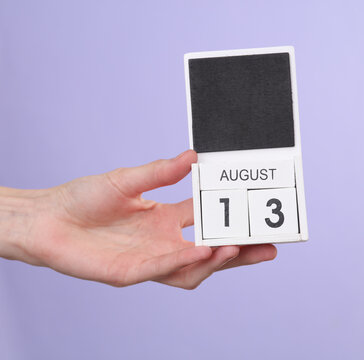 Woman's hand holds wooden calendar with date august 13 on pastel purple background