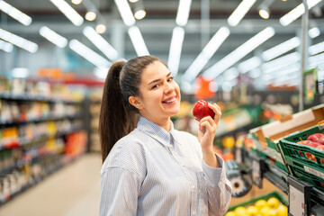 Young woman at supermarket making healthy choices by choosing organic fresh food. Charming woman with a dimple holding a red apple as a symbol of healthy eating and cheerfully smiling at the camera.