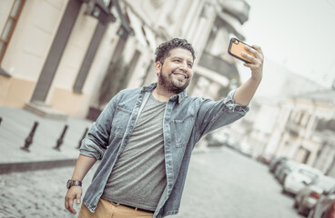 Young cheerful hispanic man in jeans jacket makes selfie on phone in european street