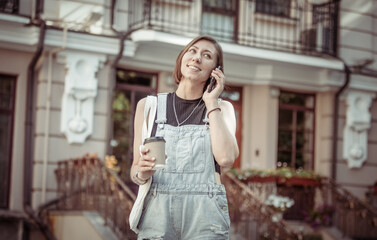 Stylish young woman in denim overalls holding cup of coffee and talking on phone in the city