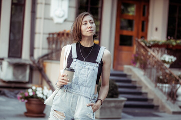 Stylish young woman in denim overalls holding a cup of coffee in the city