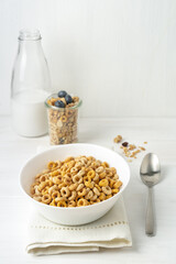 Cereal bowl, milk bottle and spoon on a beige napkin on a white table, front view.