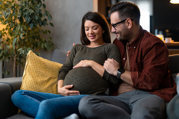 Happy couple expecting a baby. Husband caresses his pregnant wife while sitting on a couch at home.