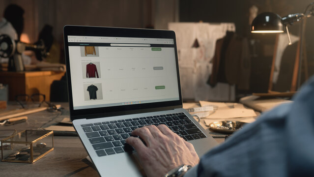 Successful man browsing personal clothing shop on laptop or watching orders statistic. Tailor sits by the table at luxury designer atelier and works with clothing range on website. Concept of fashion.