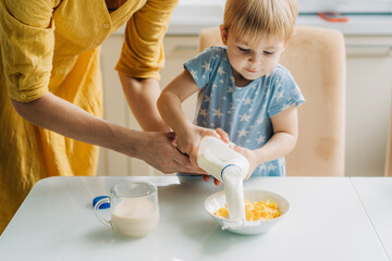 Toddler little daughter and her mother pour milk into a bowl of corn flakes.