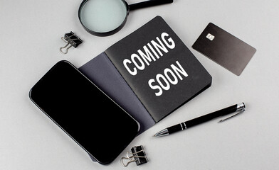 COMING SOON text written on black notebook with smartphone, magnifier and credit card