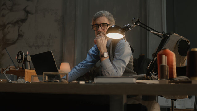 Mature tailor in black framed glasses works on new clothes collection. Man draws modern design of tailorer suit on laptop or tablet computer. Concept of fashion and craftsman job at workshop.