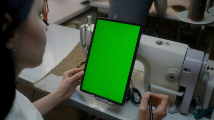 Wear designer or seamstress sits at sewing machine, watches at tablet, keeps stylus in hand and touches screen. Green screen displayed on gadget. Atelier workshop. Concept of fashion. Close up shot.