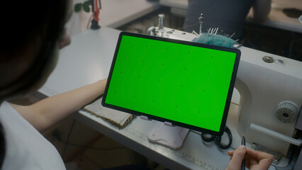 Wear designer sits at sewing machine, keeps stylus in hand, watches at tablet and touches screen. Chroma key displayed on horizontal placed device. Atelier workshop. Concept of fashion. Close up shot.