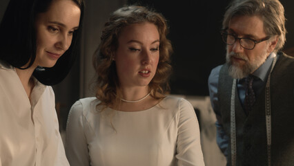 Tailors and woman talking and discussing tailoring beautiful wedding dress. Bride on fitting wedding dress in luxury designer atelier or tailoring studio. Concept of fashion, handmade and couturier.