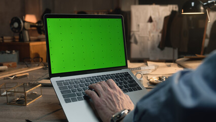 Male tailor or fashion designer creates and developes clothes on laptop in luxury atelier or tailoring workshop. Mature man sits on workplace and types at laptop with green screen. Close up view.