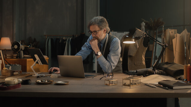 Mature couturier in black glasses works on new wear collection in atelier. Man draws modern design of tailored suit on laptop or tablet computer. Concept of fashion and craftsman job. Slow motion