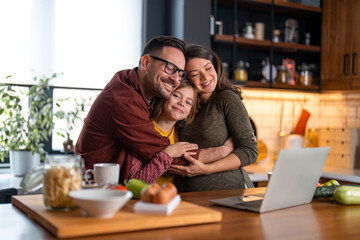 Loving family of three embracing in the kitchen. Mom and dad are daydreaming with the eyes closed while little girl is smiling and looking at the camera. They're standing at the kitchen counter.