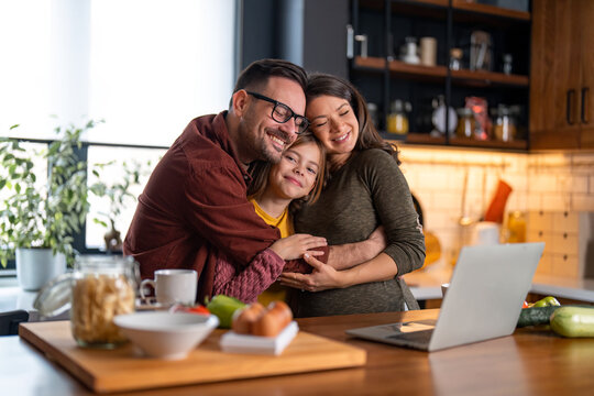 Loving Family Of Three Embracing In The Kitchen. Mom And Dad Are Daydreaming With The Eyes Closed While Little Girl Is Smiling And Looking At The Camera. They're Standing At The Kitchen Counter.