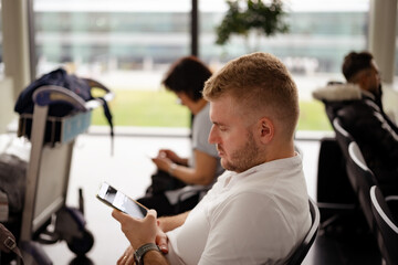 Middle-aged caucasian blond man wearing casual clothes sitting in airport, waiting for check-in, boarding,using mobile phone,texting.Travelling tourist on holidays concept.Copyspace for text.