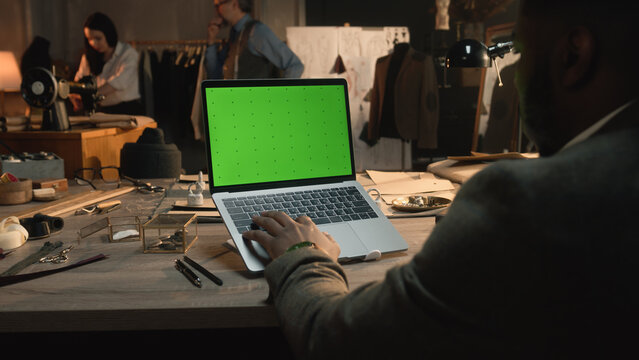 African American fashion designer sits at the table, types on keyboard, takes online orders from customers or creates clothes on laptop with green screen. Atelier view at background. Close up view.