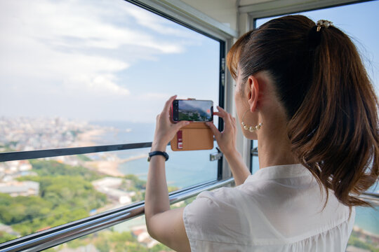 A mature Japanese woman using her mobile phone to take pictures from a cable car cabin of the city and landscape below.