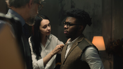 Cinematic shot of female tailor takes measurements of African American man for wedding or business suit shirt. Luxury dim atelier or tailoring studio. Concept of fashion, handmade and couturier.