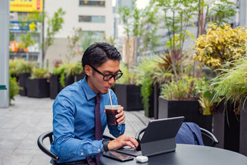 A young businessman in the city, on the move, a man seated at a cafe table outdoors, using a laptop, sipping a soft drink with a straw.
