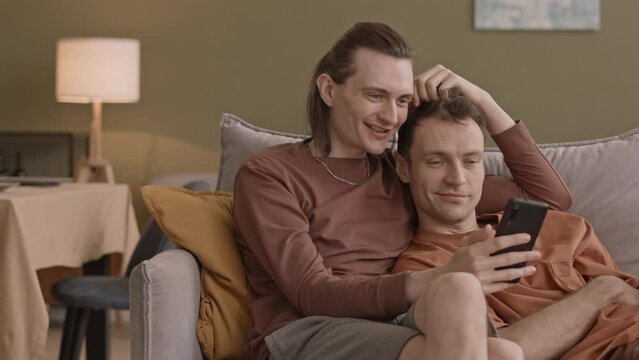 Happy peaceful gay couple spending time together at home relaxing on sofa in living room watching photos on smartphone