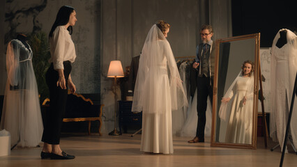 Cinematic shot of female tailor putting veil on woman in wedding dress in front of mirror. Bride on fitting wedding dress in designer atelier or wedding salon. Concept of fashion and couturier.