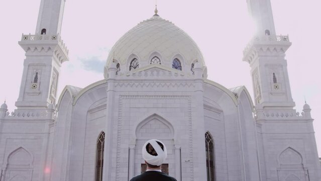 Mullah wearing turban prays in front of White Mosque and respects own religion. Man standing on square asks for good life and prays