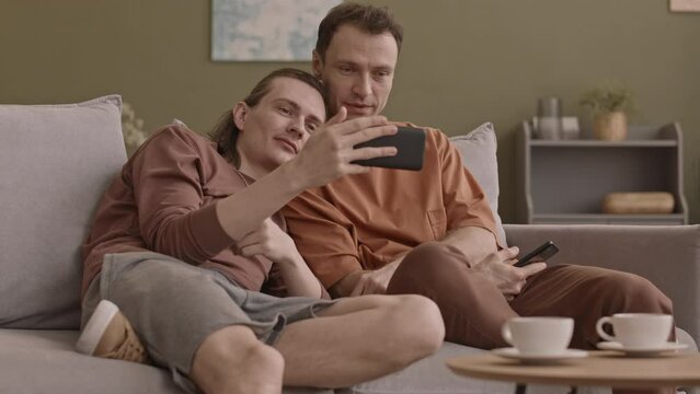 Cozy romantic gay couple resting on sofa in living room together watching video on smartphone